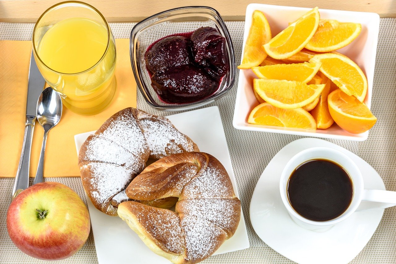 Foto de mesa posta. Em tons de amarelo, temos sucos, frutas maçã inteira e um parto com gomos de laranja, pão em formato de meia lua, geleia e uma xícara branca com café puro